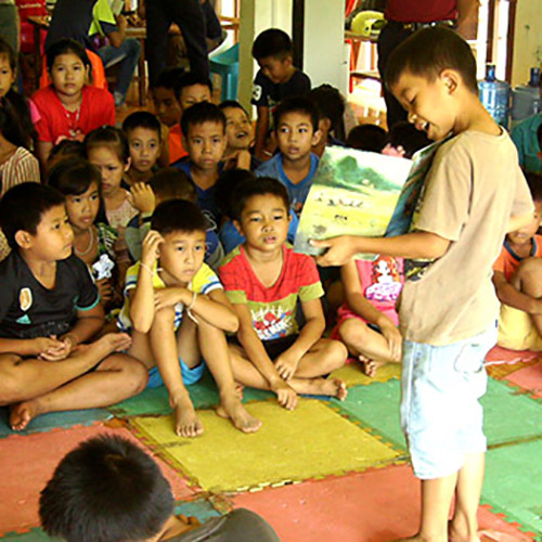 A small boy reads aloud from a book and displays the pictures to other children.