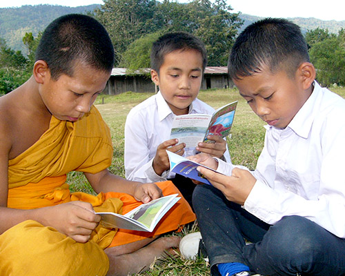 Three boys, including a novice Buddhist monk in an orange robe, sit on the grass reading books