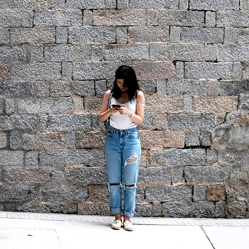 A young woman stands against a wall, looking at her phone.