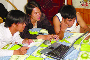 Three young Lao men, with a laptop computer on a bed, looking at a book manuscript
