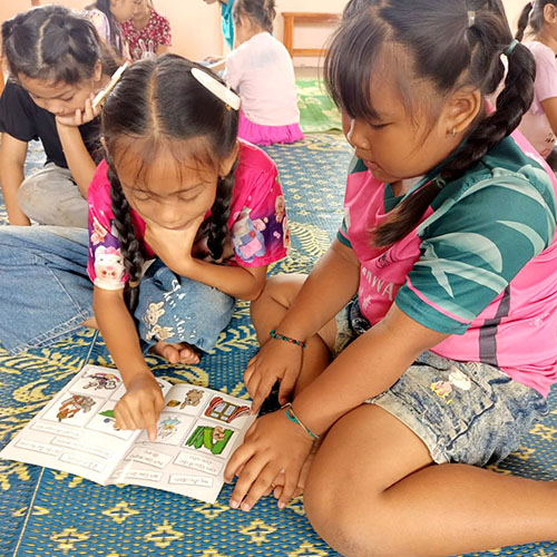One girl helps another learn to read, they are sitting on the floor with a book