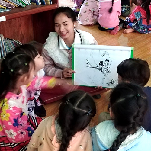 Teacher and several children sitting on the floor, she looks at a large wordless book with them.