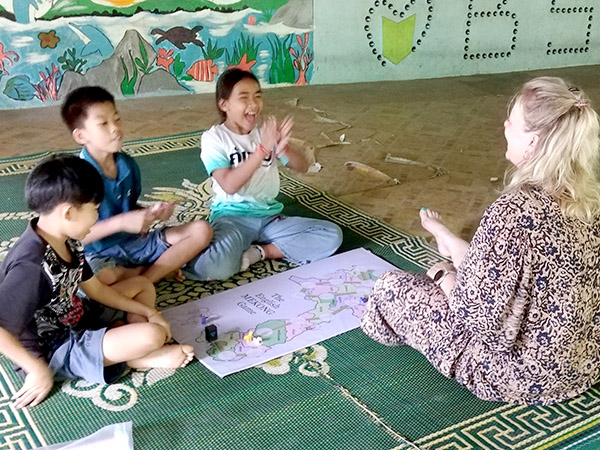 A woman and three Laotian children sit on school floor with map, playing a game to help them learn English