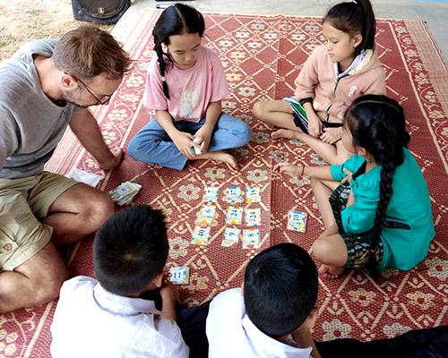Several Lao children and a visitor from Europe play a math game. Nine cards with numbers are face up.